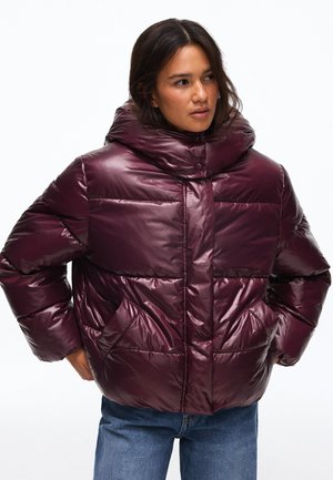 Femme portant une doudoune brillante violet foncé avec un col montant et des poches latérales, associée à un jean bleu, debout les mains dans les poches.