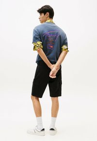 Camiseta azul con diseño gráfico en la parte de atrás, mangas a cuadros amarillos. Pantalones cortos cargo negros y zapatillas blancas con acentos negros. Calcetines blancos.