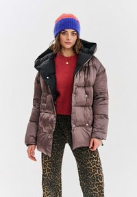 Puchowa, brązowa kurtka z czarnym kapturem, z zapięciem na guziki, noszona na czerwonym swetrze i spodniach w leopardzi wzór, w zestawie z wielokolorową wełnianą czapką.