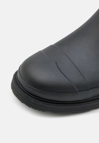 Botte en caoutchouc noire avec un bout arrondi, surface texturée et semelle épaisse à nervures. Présente des lignes horizontales sur le capuchon et une finition lisse.