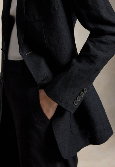 Osoba u crnom odijelu s desnom rukom u džepu, pokazujući gumbe na rukavu i dio bijele unutarnje košulje.