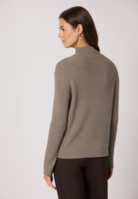 Pull en maille côtelée de couleur taupe douce, avec un col montant et des manches longues. Le design présente une coupe décontractée et un ourlet légèrement plus bas à l'arrière.