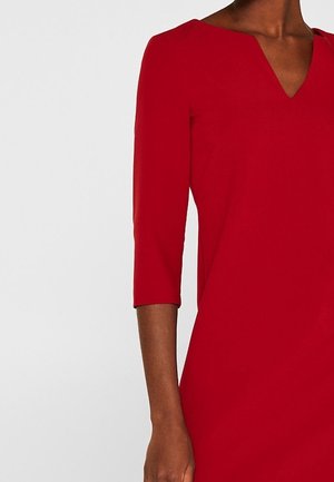 Czerwona sukienka z rękawami długości do łokcia i dekoltem w szpic, noszona przez osobę o ciemnej karnacji, pokazana od szyi do połowy uda.