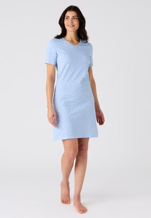 Femme debout, pieds nus, portant une robe bleu clair à manches courtes avec de petits motifs floraux blancs, regardant légèrement sur le côté et souriant.