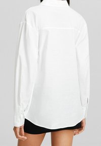 Camicia bianca a maniche lunghe con colletto, caratterizzata da una trama liscia, cucitura sulla schiena e dettagli con polsini abbottonati. L'orlo è leggermente curvato.