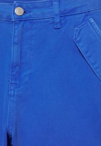 Gros plan d’un pantalon bleu vif montrant un bouton en métal, une boucle de ceinture, une poche avant gauche et une couture détaillée sur un tissu sergé.