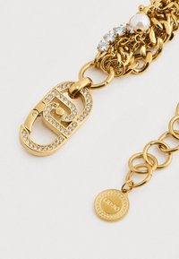 Kultaketju rannekoru, jossa on suuri solki, joka on koristeltu kirkkailla kristalleilla, helmiyksityiskohdalla ja pienellä pyöreällä merkkitarroilla.