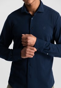 Marineblaue Langarmbluse mit durchgeknöpftem Front, weicher Textur und gesticktem Logo auf der Brust. Mit geknöpften Manschetten.