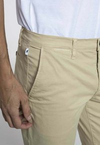 Chino beige en tissu coton lisse, coupe ajustée et poches latérales. Comprend une petite étiquette de marque sur la taille.
