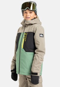 Winterjack voor kinderen in beige, groen en zwart; heeft een ritszak op de borst met een gele rits. Inclusief zwarte wanten en een bril.