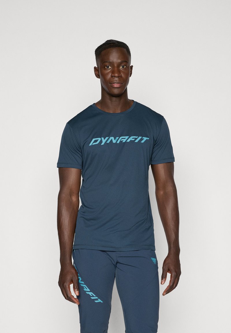 Camiseta deportiva de color azul marino con mangas cortas y cuello redondo, que presenta un logo en azul claro de "DYNAFIT" en el pecho. Textura de tela suave.