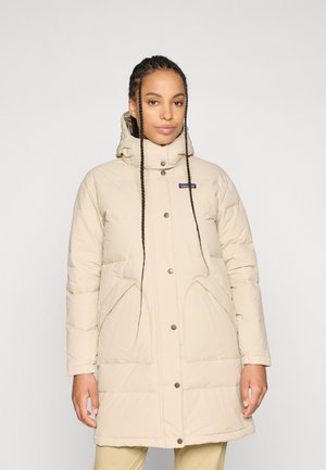 Vrouw in een beige gewatteerde lange padded jas met capuchon en drukknoopsluiting, staand tegen een effen lichte achtergrond.