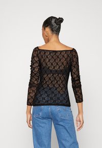 Schwarzes Spitzenoberteil mit langen Ärmeln, Rundhalsausschnitt, floralem Muster und strukturiertem Stoff. Tragen mit hellblauen, hochgeschnittenen Jeans.