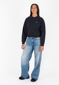 Felpa nera corta con maniche raglan e un piccolo logo sul petto, abbinata a jeans a gamba larga di colore blu chiaro. Calzature nere.