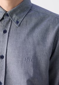 Gros plan d'une personne portant une chemise bleue à boutons avec col, avec le mot "BOSS" brodé sur la poitrine.
