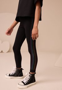 Sorte leggings med en sidebånd af farverige rhinsten, kombineret med sorte høje sneakers og en løs sort top.