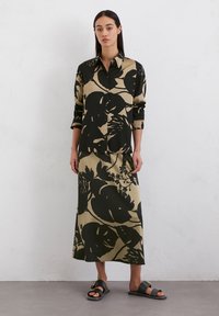 Chemise à manches longues avec boutons et jupe assortie en vert olive ornée d'un grand imprimé floral noir. Le mannequin porte des sandales plates noires avec une boucle dorée.