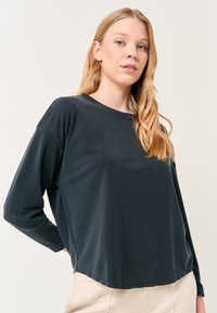 Maglietta grigia scura a maniche lunghe con vestibilità rilassata, scollatura rotonda e tessuto morbido, con orlo leggermente curvato e spalle scese.