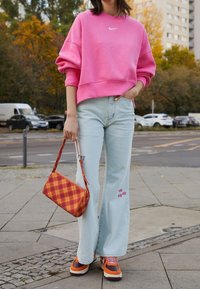 Osoba v růžové mikině Nike, světle modrých džínech s nášivkami, s oranžovo-hnědými teniskami a držením červeno-oranžové kostkované kabelky venku.