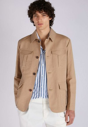 Chaqueta de lino beige con diseño de cuello, cuatro bolsillos frontales y botones marrones. Se lleva sobre una camisa a rayas azul y blanca.