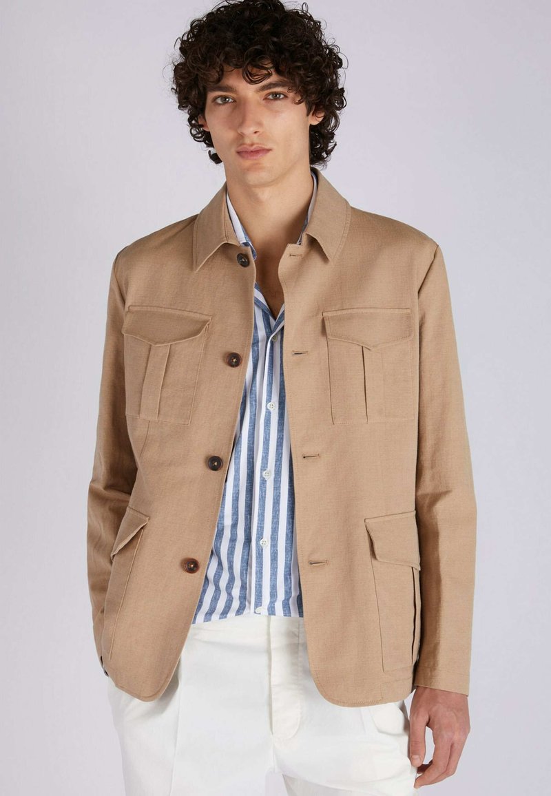 Beige Leinenjacke mit Kragen-Design, vier Fronttaschen und braunen Knöpfen. Getragen über einem blau-weißen gestreiften Hemd.