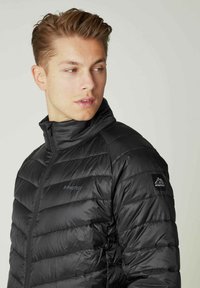 Sort pufferjakke med høj krave, kviltet tekstur og et logo på venstre arm. Lynlås foran, letvægtsmateriale.