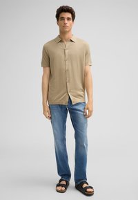 Korte mouw blouse met knoopsluiting in lichtbeige stof; gecombineerd met blauwe straight-leg jeans en zwarte sandalen; casual, ontspannen fit.