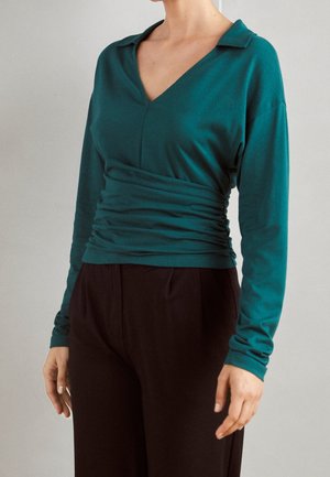 Žena v tyrkysovém topu s dlouhými rukávy a výstřihem do V, s nařaseným pasem, ve spojení s černými kalhotami s vysokým pasem na jednobarevném pozadí.