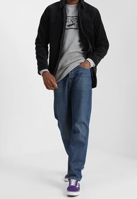 Uomo che cammina indossando una giacca nera, una felpa grigia Vans, jeans blu e sneaker Vans viola su uno sfondo neutro.