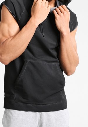 Mężczyzna w czarnej bezrękawniku z kapturem, który oburącz poprawia kaptur, z widocznymi umięśnionymi ramionami i jasnoszarymi spodniami.