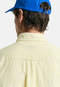 Geel gestreept shirt met een kraag en een rugplooi, gemaakt van een zachte stof. Een blauwe honkbalpet wordt gedragen, gedeeltelijk zichtbaar aan de bovenkant.