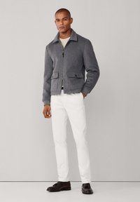 Chaqueta gris de mezcla de lana con cremallera y cuello, dos bolsillos delanteros, combinada con pantalones blancos y zapatos oscuros. Textura suave, ajuste a medida.