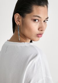 Lange, lineære øreringer med blå og klare krystaller på en sølvfargede metallbunn, iført med en hvit genser, sett fra bak.