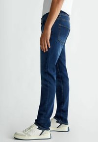 Donkerblauwe denim jeans met een slim fit, voorzien van oranje stiksels en vijf zakken. Gecombineerd met witte sneakers en een eenvoudig t-shirt.