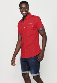 Camisa roja de manga corta con detalles de mezclilla, dos bolsillos en el pecho y detalles de botones. Combinada con pantalones cortos de mezclilla azules, con los puños doblados.