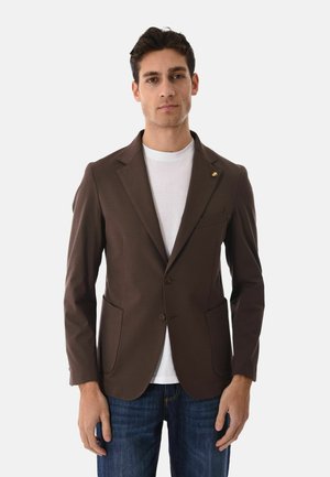 Uomo che indossa un blazer marrone sopra una t-shirt bianca e jeans blu, in piedi contro uno sfondo chiaro uniforme, volto in avanti con espressione neutra.