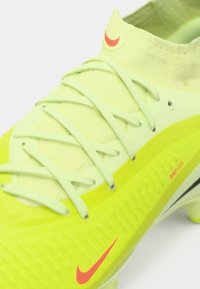 Botín de fútbol Nike amarillo brillante con cordones verde claro y logotipos de swoosh rojos en la puntera y el lateral, con un cuello de tobillo de tejido visible.