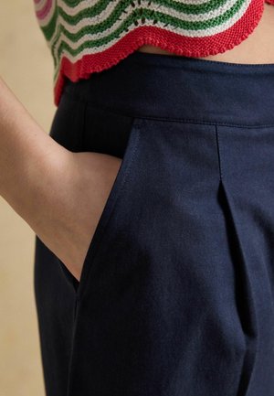 Hånd i lommen på navyblå bukser med høj talje kombineret med en flerfarvet, rød, grøn og hvid strikket top.