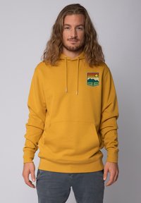 Geel hoodie met een grafisch ontwerp aan de linkerkant met bergen, zon en de tekst "GOOD VIBE." Zachte textuur, voorzak.