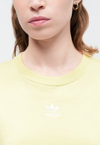 Žena v světlé žluté mikině Adidas s malým bílým logem na prsou a zlatými kruhovými náušnicemi, zobrazená od krku po rty.