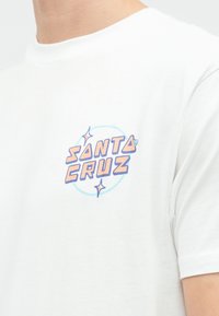 Biele tričko s krátkymi rukávmi, ktoré má na ľavej strane hrudníka pastelovo oranžové a modré logo "Santa Cruz" so hviezdami.