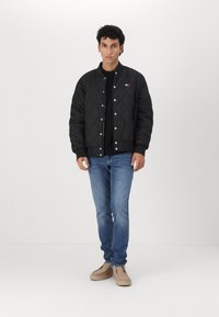 Svart quilted bomberjakke med ribbestrikket krage og trykknapper, brukt over en svart skjorte, sammen med blå jeans og beige sko.