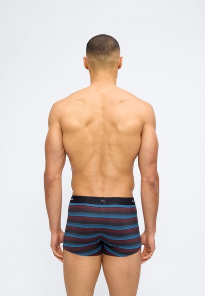 Μυώδης άνδρας με κοντά μαλλιά, γυρισμένος πλάτη, φορώντας ριγέ μποξεράκια με μπλε και βυσσινί οριζόντιες γραμμές πάνω σε απλό ανοιχτό φόντο.