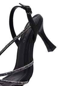 Schwarze High-Heelsandale mit mehreren verzierten Riemen, ausgestattet mit einem glänzenden silbernen Akzent, einem Knöchelverschluss und einem schmalen Stiletto-Absatz.
