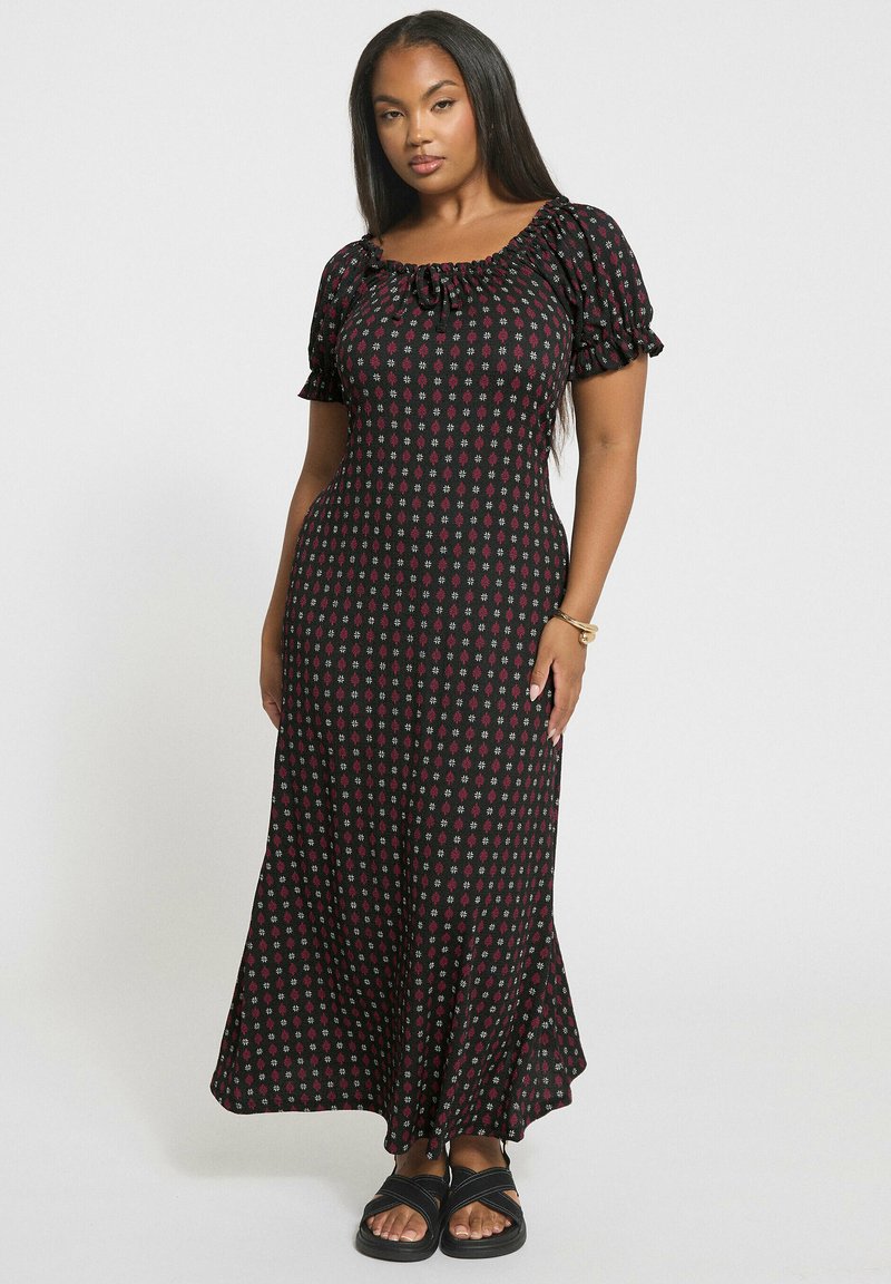 Kvinna med långt mörkt hår som bär en svart maxiklänning med röd och vit blommönster, puffärmar och svarta sandaler, stående mot vit bakgrund.