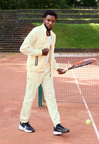 Light yellow sports tracksuit with a zip-up jacket and elastic waistband pants. Black sneakers and a tennis racket in hand, on a clay court.