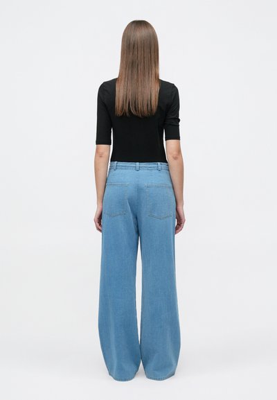 Γυναίκα με ίσια καστανή τρίχα στέκεται με την πλάτη, φορώντας μαύρο τοπ με μισά μανίκια και ανοιχτόχρωμα μπλε τζιν παντελόνια με ψηλή μέση και φαρδιά γραμμή.
