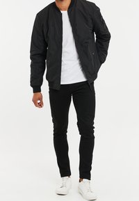 Giacca bomber nera con chiusura frontale a zip, polsini a coste e tasche laterali; indossata sopra una t-shirt bianca, abbinata a pantaloni neri slim-fit e sneakers bianche.