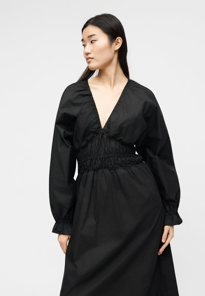 Γυναίκα που φοράει μαύρο φόρεμα με μακριά μανίκια, βαθύ V-ντεκολτέ και σφηκοφωλιά στη μέση, κοιτάζοντας προς τα αριστερά με λευκό απλό φόντο.