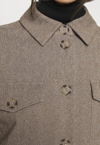 Brun skjortjacka med fiskbensmönster, krage, två bröstfickor och fyra stora knappar i dämpad ton. Nära bild av den texturerade tyget.